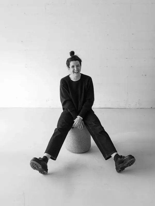 Portrait professionnel en noir et blanc de Sacha, assise sur un pouf dans un studio aux murs de brique blanche