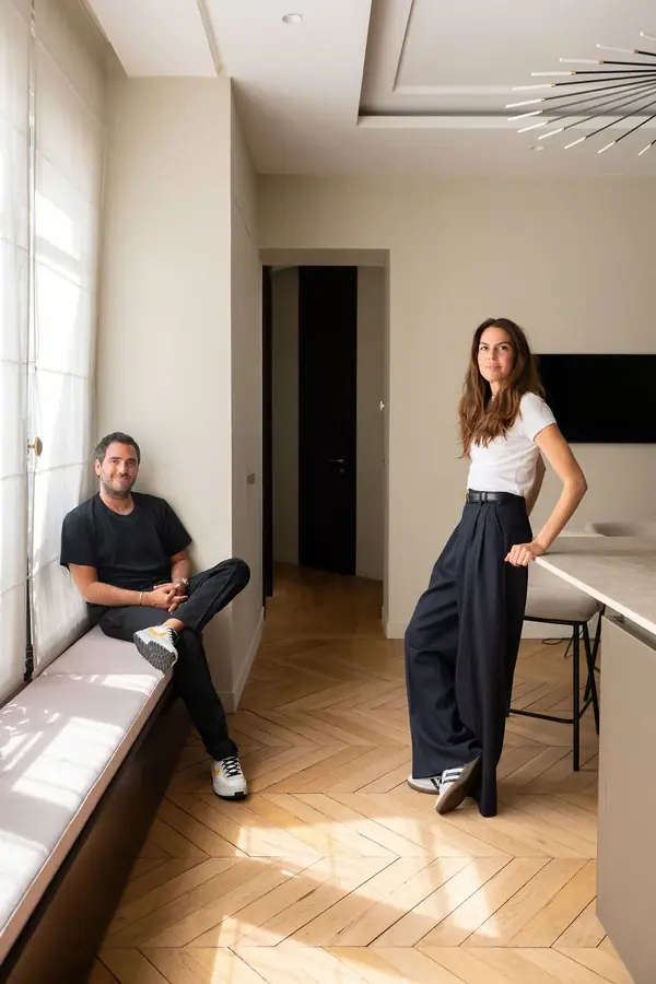 Portrait professionnel d'un duo dans un appartement lumineux, lui assis sur une banquette, elle debout sur un parquet