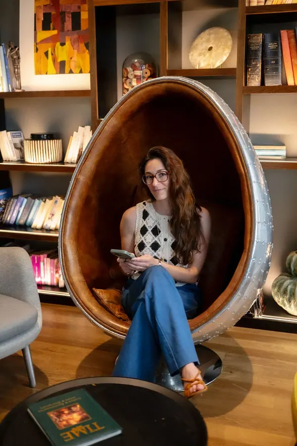 Portrait professionnel de Bénédicte assise dans un fauteuil œuf, livre en main, devant une bibliothèque aux tons chaleureux