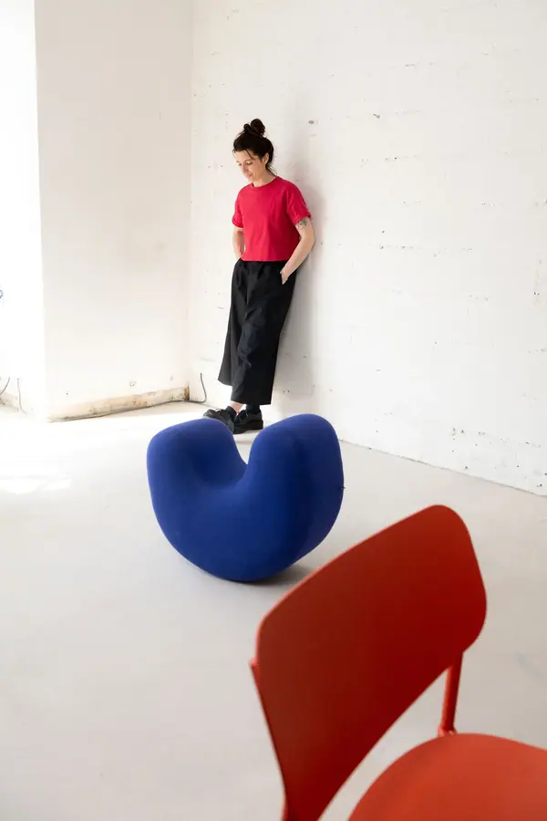 Portrait de Sacha en tee-shirt rouge, adossée à un mur de briques blanches, près d'une assise bleue et d'une chaise rouge