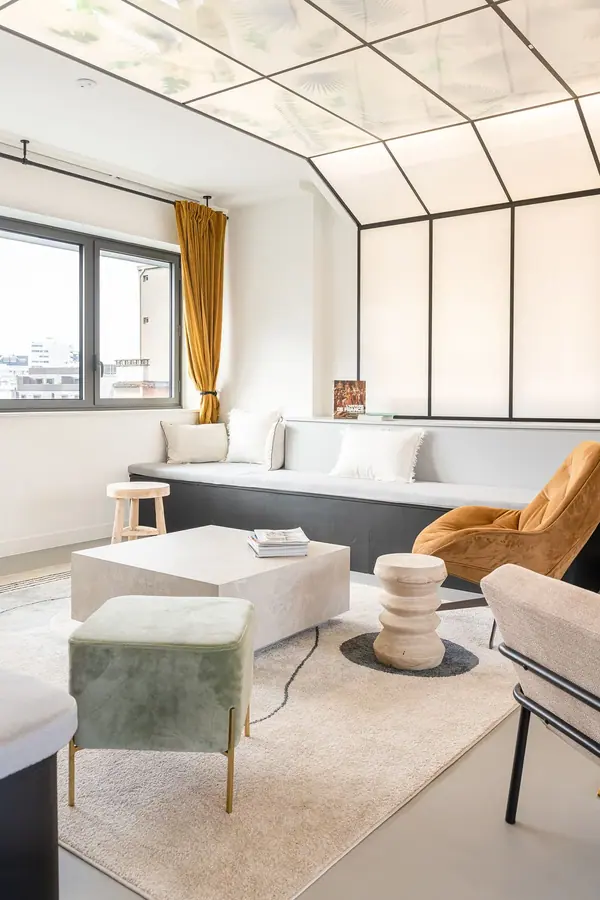 Salon des bureaux Newen Studios par Yemanja : verrière à cadres noirs, rideaux moutarde et fauteuil en rotin, lumière douce