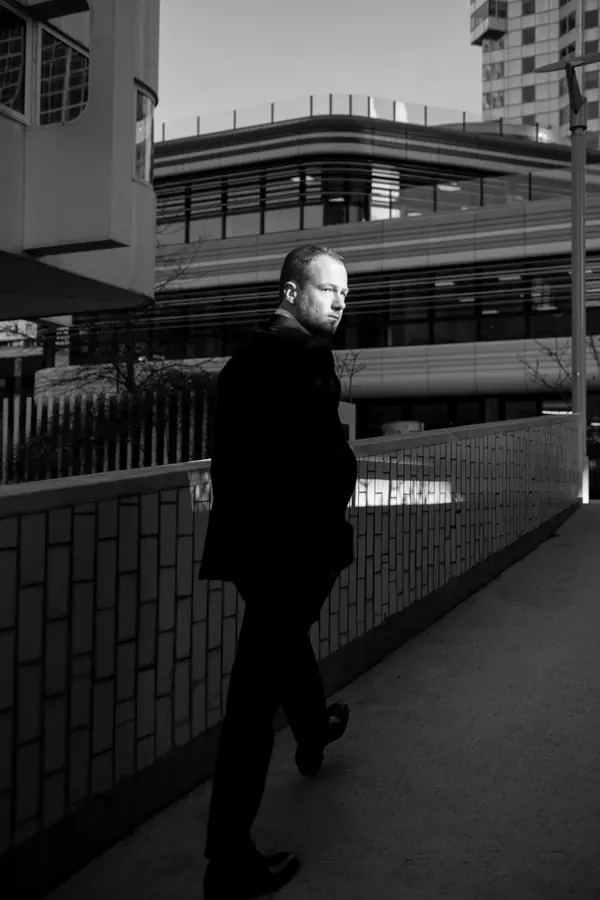 Portrait corporate noir et blanc d'un homme en veste sombre marchant sur une passerelle devant des immeubles modernes