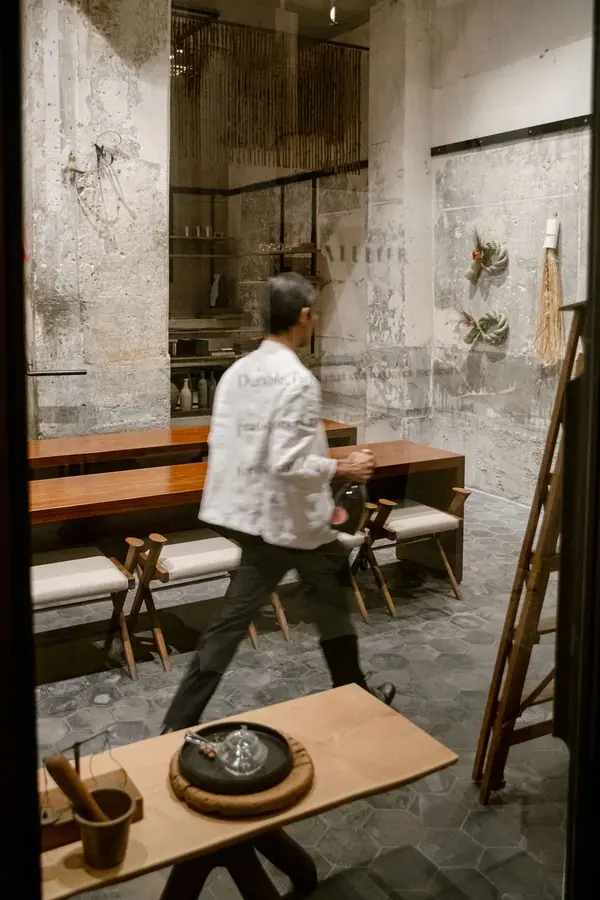 Reportage photo chez OGATA Paris, un chef en veste blanche traverse une salle aux murs de pierre et tables en bois