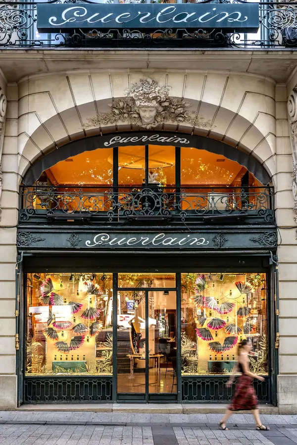 Façade historique de la boutique Guerlain sur les Champs-Élysées, vitrines illustrées et passante en robe rouge