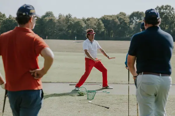 Coach de golf en pantalon rouge donnant un cours au practice à des participants d'une journée d'entreprise, vus de dos au premier plan
