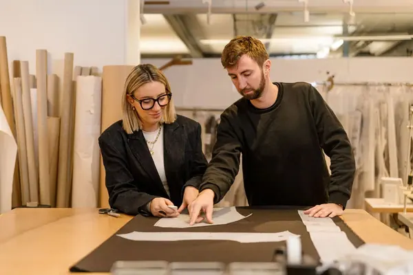 Portrait professionnel de deux apprentis LVMH penchés sur un patron en papier sur une table d'atelier, lumière douce